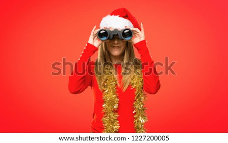 Blonde woman dressed up for christmas holidays and looking for something in the distance with binoculars on red background