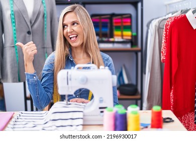 Blonde Woman Dressmaker Designer Using Sew Machine Smiling With Happy Face Looking And Pointing To The Side With Thumb Up. 