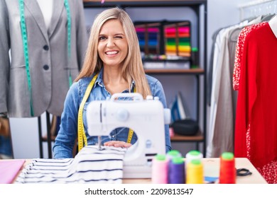 Blonde Woman Dressmaker Designer Using Sew Machine Winking Looking At The Camera With Sexy Expression, Cheerful And Happy Face. 