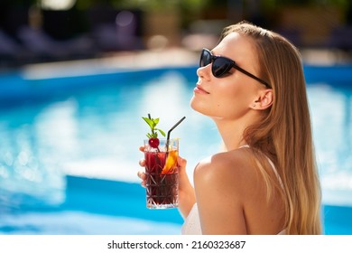 Blonde Woman Drinks Refreshing Cocktail Sunbathing And Sitting Near Swimming Pool At Tropical Spa. Female In Sunglasses Enjoys A Drink On Poolside. Girl Chilling In Tropical Resort On Vacation.