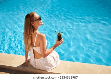 Blonde woman drinks refreshing cocktail sunbathing and sitting near swimming pool at tropical spa. Female in sunglasses enjoys a drink on poolside. Girl chilling in tropical resort on vacation. - Powered by Shutterstock