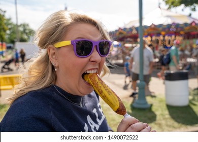 Blonde woman eats a pronto pup corn dog covered in mustard at an outdoor fair