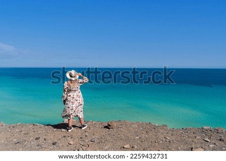 Blonde woman in a flowery dress with her back turned holding a straw hat in her hand in front of the blue sky and turquoise sea on the coast of the touristic Fuerteventura in the Canary Islands.