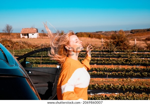 Blonde Woman with flying hair on the wind standing by\
the car at countryside farm. Travelling across the country. Copy\
space 