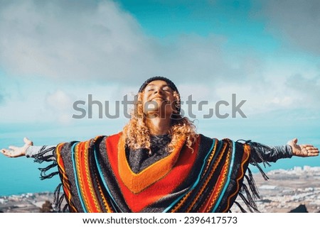 Blonde woman in hat and colorful poncho relaxing with open arms in blue winter sky. People's freedom style. People  freedom style. Traveler outstretching arms outdoor leisure activity