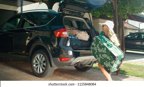 Blonde Woman Having Trouble With Lifting A Heavy Suitcase Into The Large Black Car Parked In The Sunny Driveway Of A House In The Tranquil Suburbs. Happy Girl Getting Ready To Go On A Fun Road Trip.