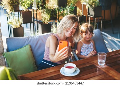 Blonde Woman And Her Little Daughter Using Smartphone For Video Chat In The Cafe. Lifestyle, Technology And Communication Concept.