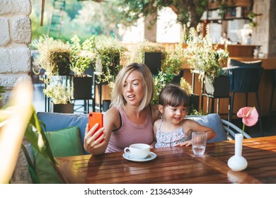 Blonde Woman And Her Little Daughter Using Smartphone For Video Chat In The Cafe. Lifestyle, Technology And Communication Concept.