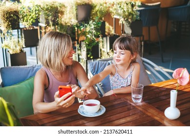 Blonde Woman And Her Little Daughter Using Smartphone For Video Chat In The Cafe. Lifestyle, Technology And Communication Concept.