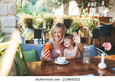 Blonde Woman And Her Little Daughter Using Smartphone For Video Chat In The Cafe. Lifestyle, Technology And Communication Concept.