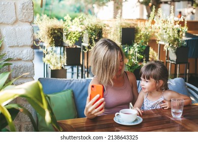 Blonde Woman And Her Little Daughter Using Smartphone For Video Chat In The Cafe. Lifestyle, Technology And Communication Concept.