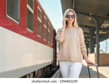 Blonde Woman With Her Luggage Go And Speak In Her Mobile Near The Red Train On The Peron Os Rail Station Under Sun Light At Sunny Day.