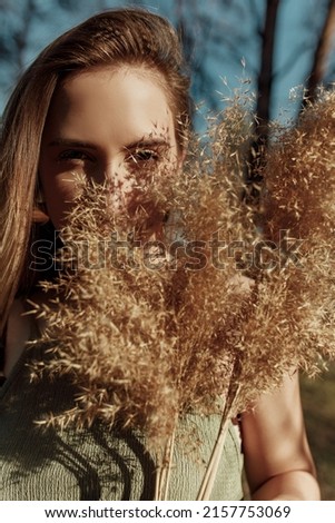 Blonde woman hiding her face by dry boho grass and part of the face is covered by pampas grass. Female model posing outdoor on hard sun light and looking at camera