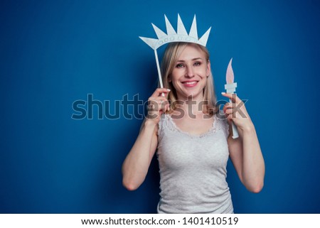 blonde woman holding American flag with paper crown and torch Statue of liberty on a blue background in the studio .4th july independence day celebration concept and learning English language