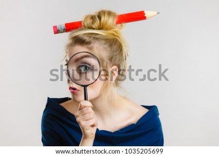 Blonde woman holding magnifying glass investigating something and looking closely, trying to find solution having big pencil in hair