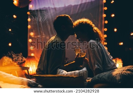 Blonde woman and husband watch movie on large screen hung on fence while lie on rest place with plaid and pillows in cottage yard backside view.