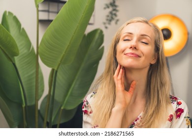 Blonde Woman In Living Room Next To Tall Plant Looking Relaxed And Smiling Holding Hand To Her Neck Delicately. Indoor Shot. High Quality Photo