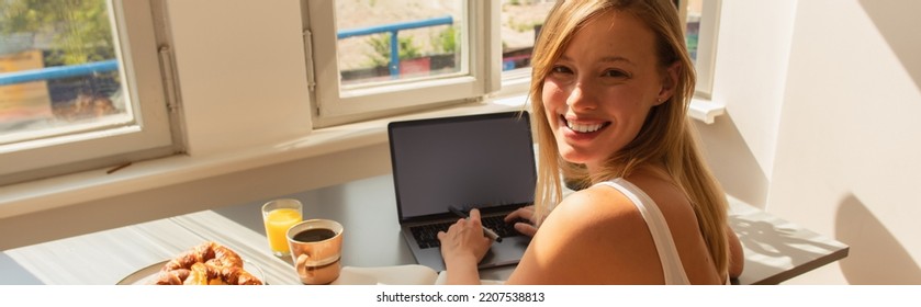 Blonde Woman Looking At Camera While Using Laptop Near Breakfast At Home, Banner