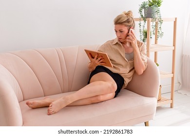 Blonde Woman, Looking At A Digital Tablet And Talking On A Mobile Phone Lying On A Beige Sofa