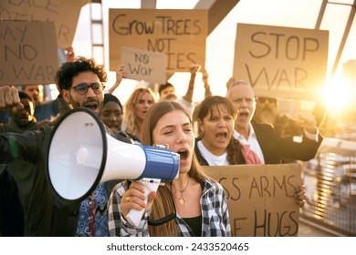 Blonde woman with megaphone in protest with diverse group activist people demonstrate together shouting slogans against wars in world holding banners for peace, love, hugs and not armies. Lens flare  - Powered by Shutterstock