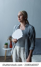 Blonde Woman With Menopause Suffering From Heat And Holding Fan In Bedroom