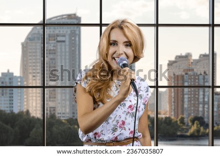 Blonde woman with microphone singing or giving a speech. Indoor window in the background.