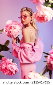 Blonde Frau in rosafarbenem Rippenspringanzug und Sonnenbrille mit rosafarbenem Peony um. Gesunde Haut und rosafarbene Schminke. rosafarbener und violetter Hintergrund. Studioaufnahme.                               