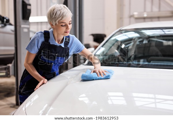 Blonde woman polish the white car hood with blue\
rag after waxing the car for shiny look ,manual car wash and hand\
wash by woman in auto service. side view on hardworking woman in\
overalls