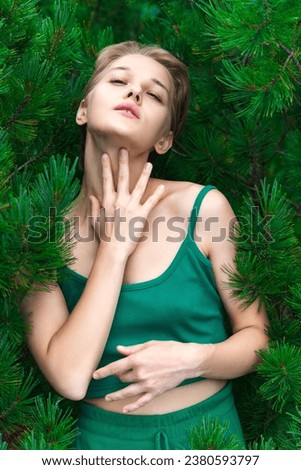 Blonde woman posing against backdrop of an evergreen shrub of Siberian Dwarf Pine. Beauty Caucasian ethnicity female looking at camera, dressed in green crop top. Part of series