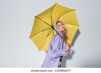 Blonde Woman Purple Rain Jacket Standing Under Yellow Umbrella On Grey