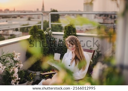 Blonde woman reading a book on urban rooftop garden. Girl sitting in chaise lounge and relaxing with cup of tee or coffee and paper book. Female enjoying life on cozy terrace with flowers and plants.