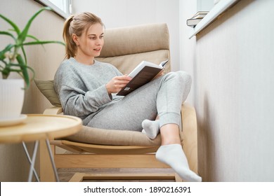Blonde woman reading book while sitting on beige armchair in apartment - Shutterstock ID 1890216391