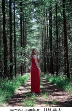 Blonde woman in red dress on forest path looking over shoulder.