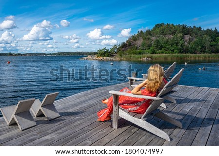 A blonde woman in a red long dress on the seaside bay. A beautiful girl is resting outdoors sitting in a chair on a wooden pier. The female in red relaxing and looking at the water. Summer vacation.