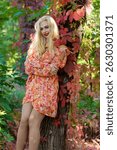 A blonde woman in a red-orange dress with long sleeves poses in the park, leaning against a tree with colorful autumn leaves.