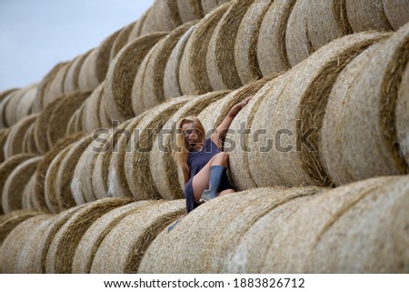 Blonde woman with rubber boots posing in a field on a hay roll