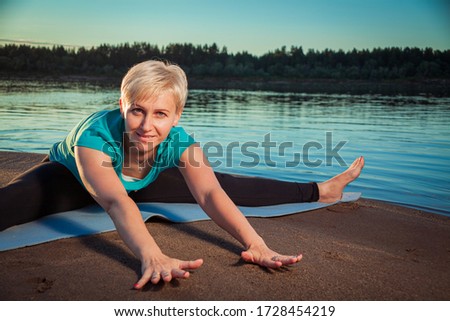 A blonde woman with a short haircut on a sandy beach near the water on a blue rug does stretching exercises, stretches her arms to the feet