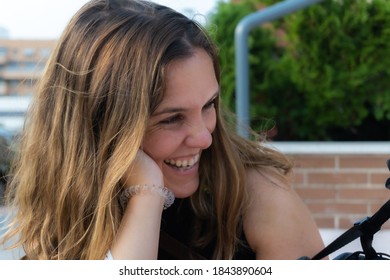 Blonde Woman Smiling Sideways With Her Head Stuck In Her Hand, In The Background A Brick Wall And A Tree