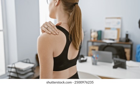 A blonde woman in a sports bra experiences shoulder pain at her office, exemplifying workplace discomfort with a blurry interior background. - Powered by Shutterstock