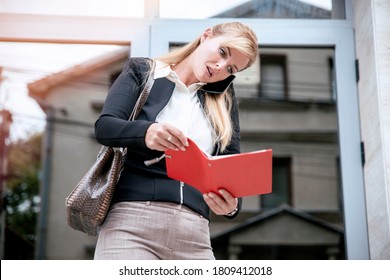 Blonde Woman Standing In Front Of Business Building Entrance And Using Mobile Phone. Young Businesswoman Busy Working Online And Talking On Her Cellphone