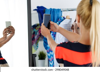 Blonde woman standing near wardrobe rack full of clothes and mirror making selfie with cellphone in new dress. Shopping and consumerism or stylist concept. Nothing to wear and hard to decide concept