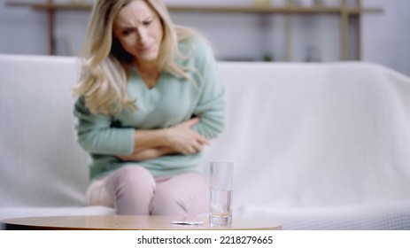 Blonde Woman Suffering From Stomach Pain Near Glass Of Water And Bottle With Pills On Coffee Table 