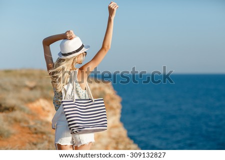 Blonde woman in summer hat and beach bag standing on the edge of the rock by the sea. Sea view. Freedom. Wind. Summer. Vacation. Yoga. looking to a sky and sea. Looking forward