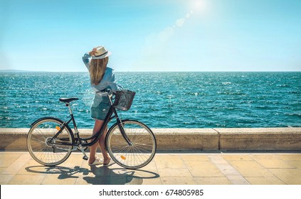 Blonde woman in summer hat with her bicycle walking coastline by the sea beach. Sea view. Freedom. Wind. Summer. Beach. Vacation. Looking to a sky and sea. Looking forward.