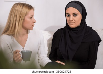 Blonde Woman Supporting Sad Muslim In Hijab, Sitting On Sofa