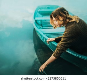 A blonde woman in a sweater is sitting in a boat floating in a lake and wants to touch the blue water surface with her hand, in which the sky and clouds are reflected. Traveling by water transport. - Powered by Shutterstock