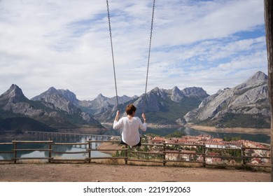 Blonde Woman Swinging On A Giant Wooden Swing, In Front Of The Amazing Mountainous Landscape Of The Riaño Damming In León, Spain.