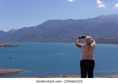 Blonde Woman Taking A Picture In A Beautiful Scenery With A Lake And Mountains On A Cloudy Day.