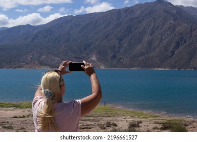 Blonde Woman Taking A Picture In A Beautiful Scenery With A Lake And Mountains On A Cloudy Day.