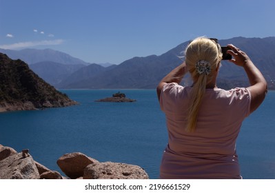 Blonde Woman Taking A Picture In A Beautiful Scenery With A Lake And Mountains On A Cloudy Day.